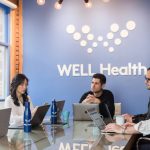 a group of five people is meeting in a board room with the logo of the Well Health company on the wall