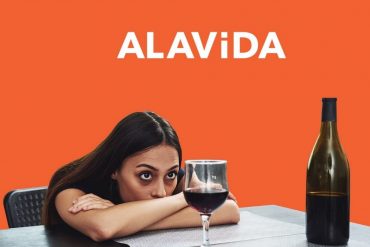 A woman with her arms folded on the table, sitting across an open bottle of wine and a glass of wine half-full. The ALAViDA logo is in white text and an orange background.