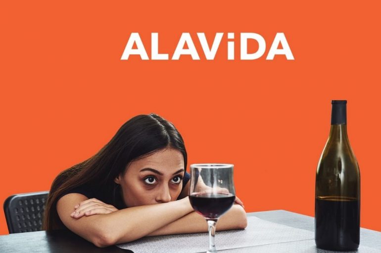 A woman with her arms folded on the table sitting across an open bottle of wine and a glass of wine half full The ALAViDA logo is in white text and an orange background
