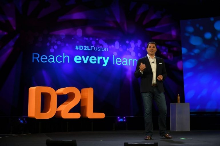 D2L founder John Baker standing on a stage with the D2L logo behind him