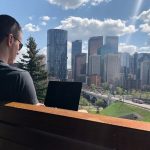 Man sitting on a bench, working on his laptop with a view of Calgary, Alberta in front of him