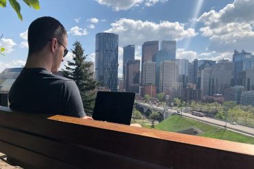 Man sitting on a bench, working on his laptop with a view of Calgary, Alberta in front of him