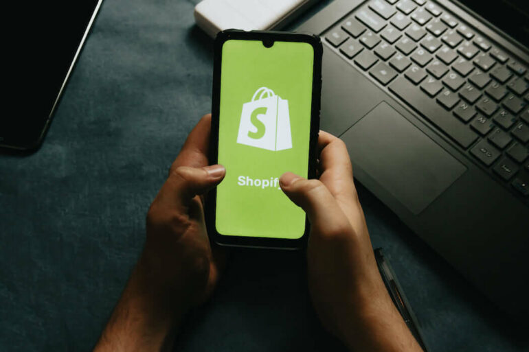 A pair of hands holding a mobile phone which has the Shopify logo on it
