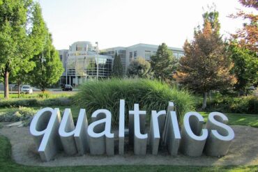 A set of three-dimensional letters coming out the ground to spell "Qualtrics" amongst some brush in front of an office-style building