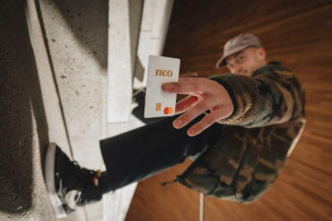 a man in skateboard shoes leans sideways and flashes a Neo mastercard at the camera. it is in focus in the foreground and he is out of focus in the background