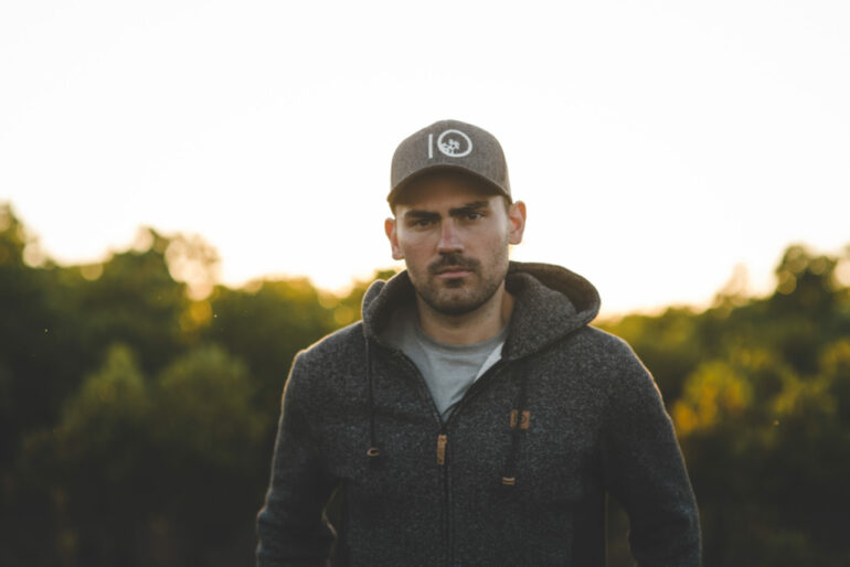 outdoor portrait of Tentree and Veritree CEO Derrick Emsley. he wears a branded baseball hat and hoodie with out of focus forest in the background