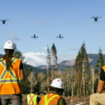 three workers in visibility vests observe as drones take off over burn landscape with mountains and forest in the background