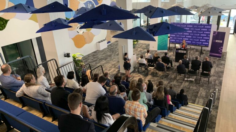 people sit in edmonton unlimited's event space watching a female speaker. cool lighting fixtures that look like umbrellas are overhead. there's a steeply tiered seating area and about 60-70 people in attendance