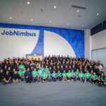 A group photo of the JobNimbus and SumoQuote employees in front of a wall with JobNimbus branding. Some are wearing SumoQuote-style mint green shirts, while many are wearing dark blue.